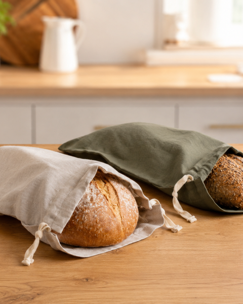 Two medium-sized linen bread bags lying side-by-side on a wooden kitchen counter. The bag on the left is a natural beige color, and the one on the right is an olive green shade. Both contain round loaves of bread with the crusty tops slightly exposed from the drawstring openings.