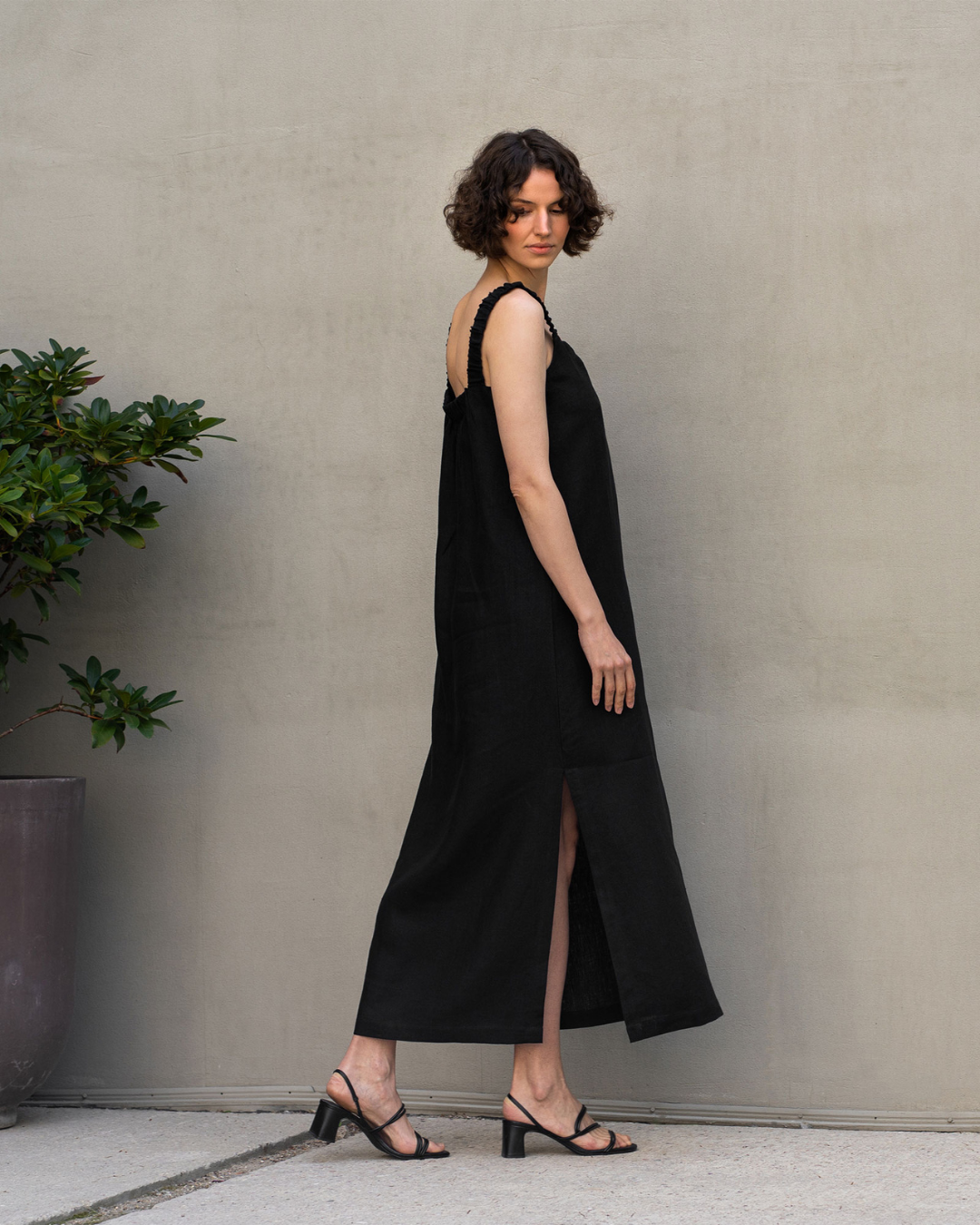 A woman wearing a black sleeveless linen dress with a side slit, photographed in profile against a light neutral wall. The minimalist summer look highlights the natural texture of linen and the relaxed, flowing silhouette of the dress.