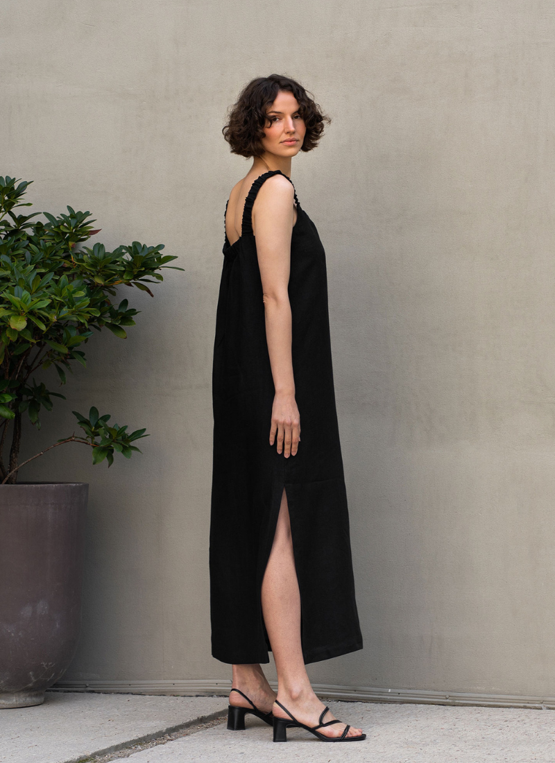A woman wearing a black sleeveless linen dress with a side slit, photographed in profile against a light neutral wall. The minimalist summer look highlights the natural texture of linen and the relaxed, flowing silhouette of the dress.