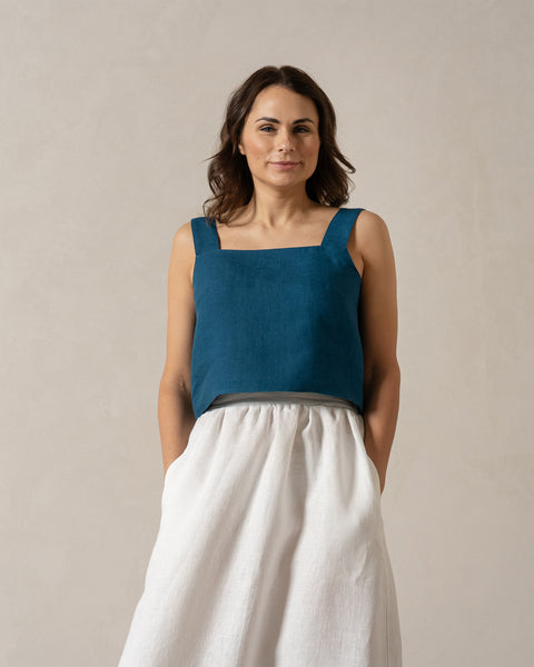 A woman with medium-toned skin and dark hair looks directly at the camera with a neutral expression. She is wearing a teal, sleeveless square-neck linen top and a pure white, menique
 linen skirt, with her hands in the skirt's pockets. The background is a plain, light beige color.
