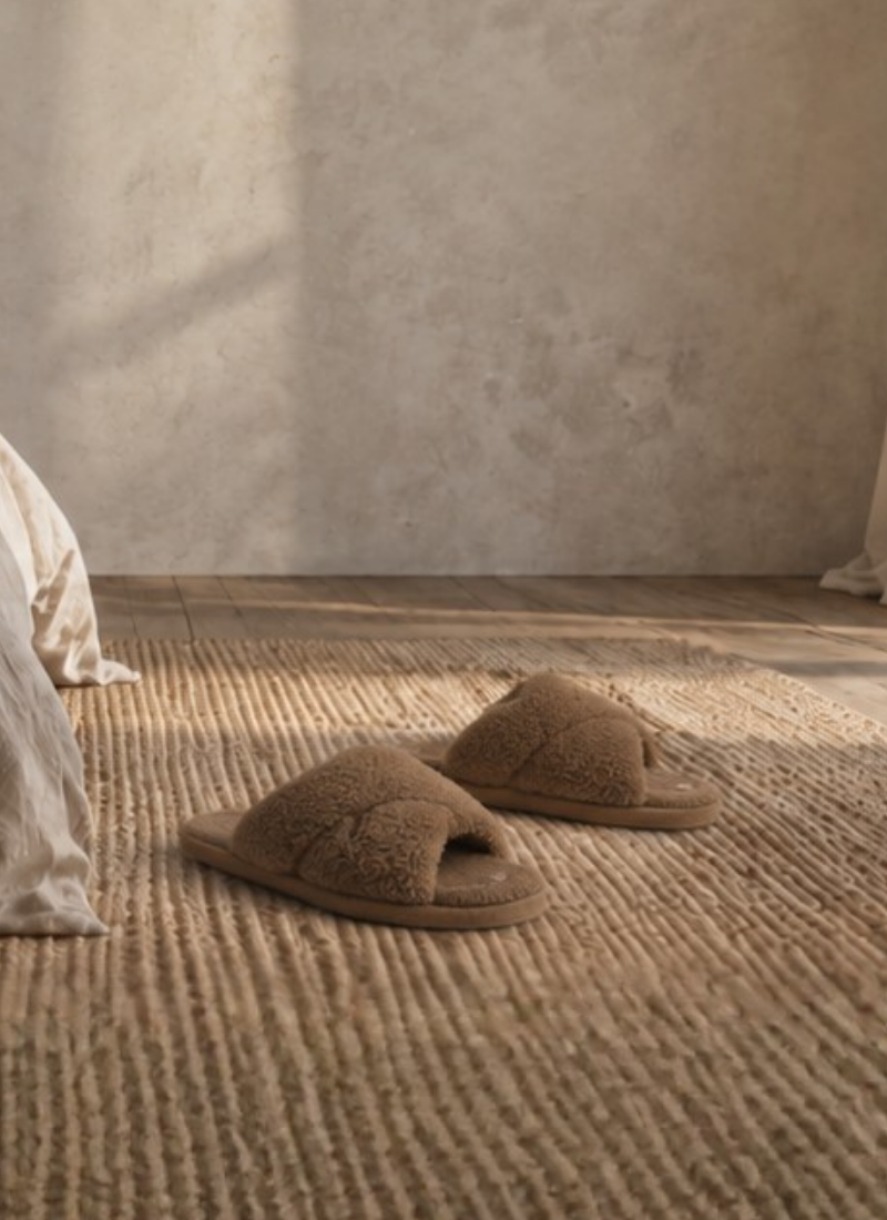 A pair of fluffy brown criss-cross slippers resting on a textured woven rug next to a bed with rumpled neutral linen sheets in a sunlit room.