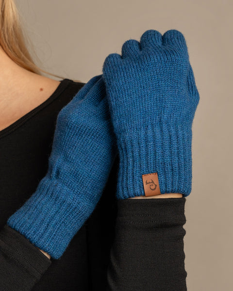A close-up shot of a person wearing menique brand vibrant blue, ribbed knit gloves. The person is wearing a dark long-sleeved top, with the right hand holding the left wrist, showcasing the gloves' fit and the small, rectangular tan leather menique tag on the cuff of the left glove. The background is a solid, neutral gray.