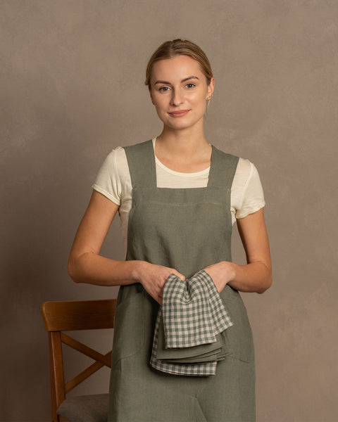 A woman holding two linen dish towels, one in stone green and the other in green check color, layered on top of one another in her hands. She is dressed in a stone green apron and a light short shirt. Behind her is a chair. 