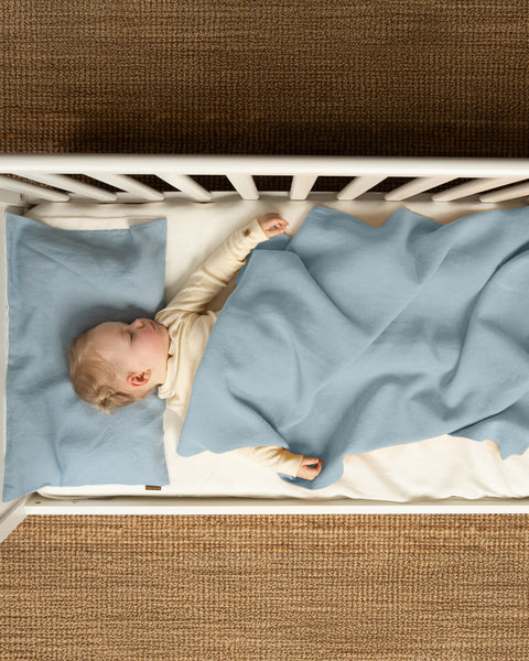 Baby sleeping in a white crib under a light blue linen blanket, on a natural brown woven rug