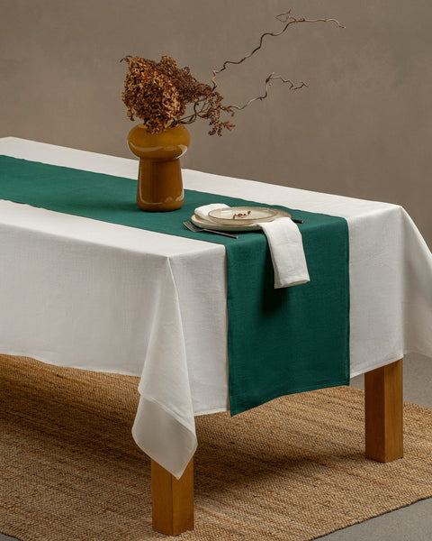 Het menique linnen tafellaken in puur witte kleur en de tafelloper in donkergroene kleur, die over een houten tafel hangt, met een glazen vaas en een theekopje met een schoteltje erop.