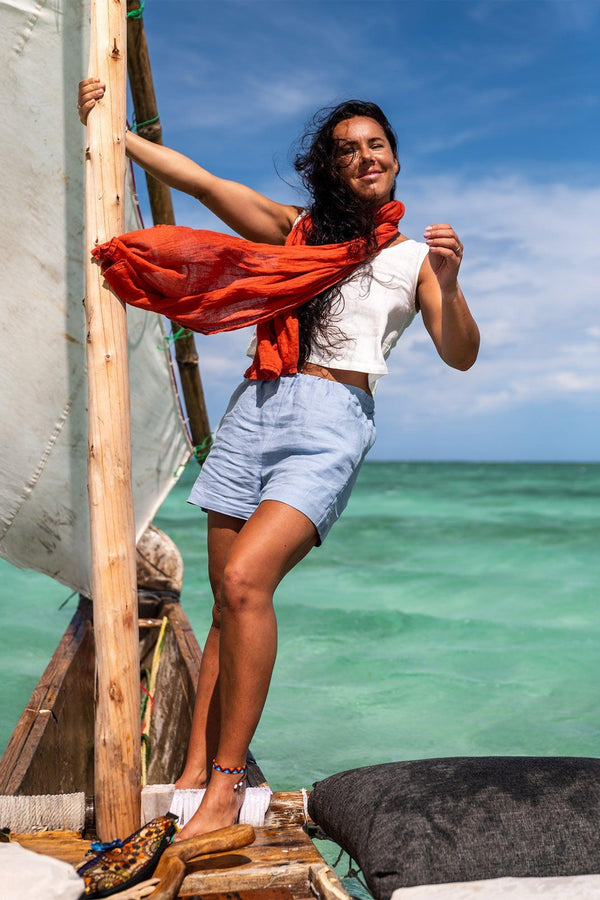 Een vrouw staat op een rustieke houten boot met een wit zeil tegen een helderblauwe lucht en turquoise water. Ze draagt een wit mouwloos hemd, lichtblauwe korte broek en een oranje sjaal. De boot heeft een wit kussen en een donkergrijs kussen.