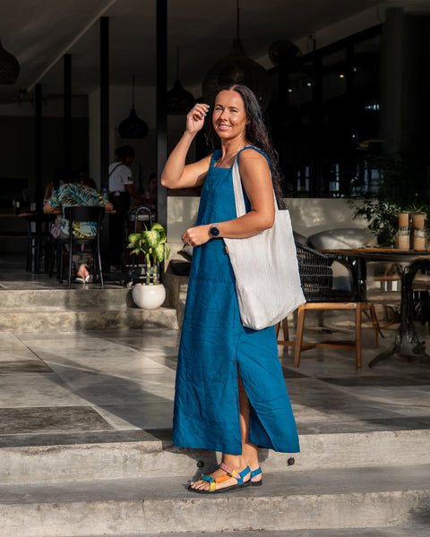 A smiling woman with long, dark hair is captured in a full outdoor shot, wearing a cobalt blue linen sundress with a square neckline, thick straps, and a side slit revealing colorful sandals. She has a menique linen tote bag slung over her shoulder and is touching her hair with one hand. The background features a building with open doorways showing interior tables, chairs, and a few people seated. Stone steps lead up to the building's entrance, and potted plants add touches of greenery.