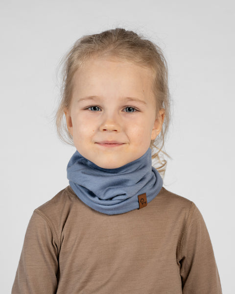 A close-up studio portrait of a young girl looking directly at the camera with a gentle smile against a plain light grey background. She has blonde hair pulled back and is wearing a brown long-sleeved top with a light blue neck gaiter bunched around her neck, featuring a small brown logo tag.