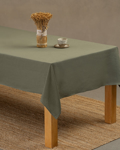 Het menique linnen tafelkleed in steengroene kleur op een houten tafel. Op het tafelkleed staat een vaas gemaakt van natuurlijke stroken en een kleien beker. Onder de tafel ligt een kleed.