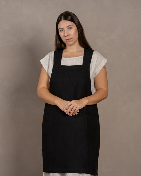 Een jonge vrouw met donker haar staat voor een studioachtergrond, met haar handen voor zich samengevouwen. Ze draagt een linnen jurk met daarover een linnen Japanse schort met kruisbanden in puur zwarte kleur, maat L/XL.