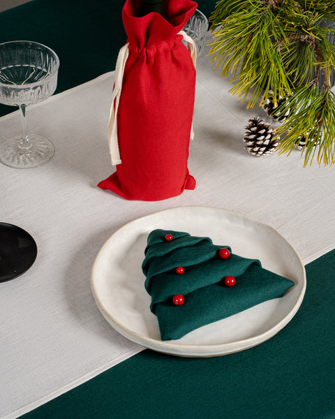 A Christmas table with a plate on top, with the menique linen napkin in dark green color folded into a christmas tree shape.