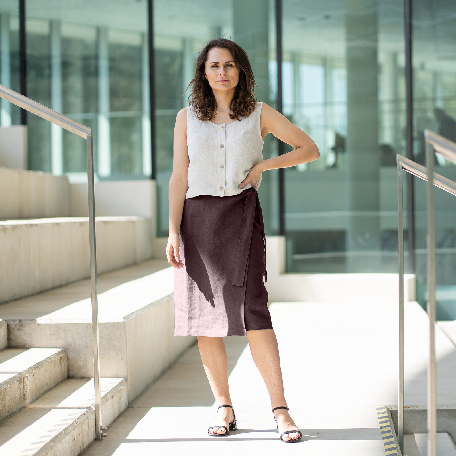 A woman with brown hair, wearing a natural linen button-front sleeveless top and a stone green wrap skirt, poses on outdoor concrete stairs.

