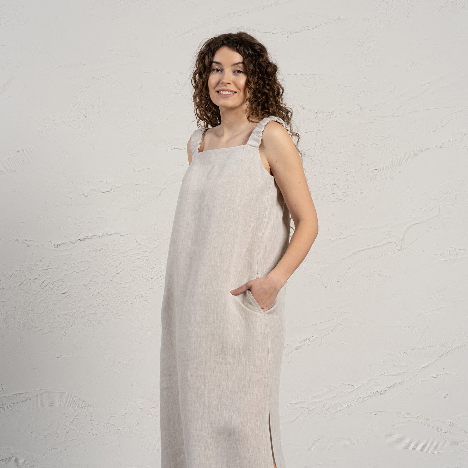 Een lichtgetinte vrouw met lang, donker krullend haar staat met een zachte glimlach naar de camera gericht. Ze draagt een mouwloze menique maxi-jurk met een vierkante halslijn en zijsplitten, en zwarte sandalen. Haar handen zitten in de zakken van de jurk, en ze staat op een lichtgrijze vloer tegen een gestructureerde gebroken witte muur.