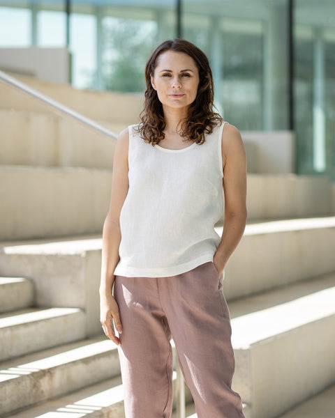A woman with brown, shoulder-length hair, wearing a white sleeveless top and faded rose trousers, stands on concrete stairs inside a building with glass walls.