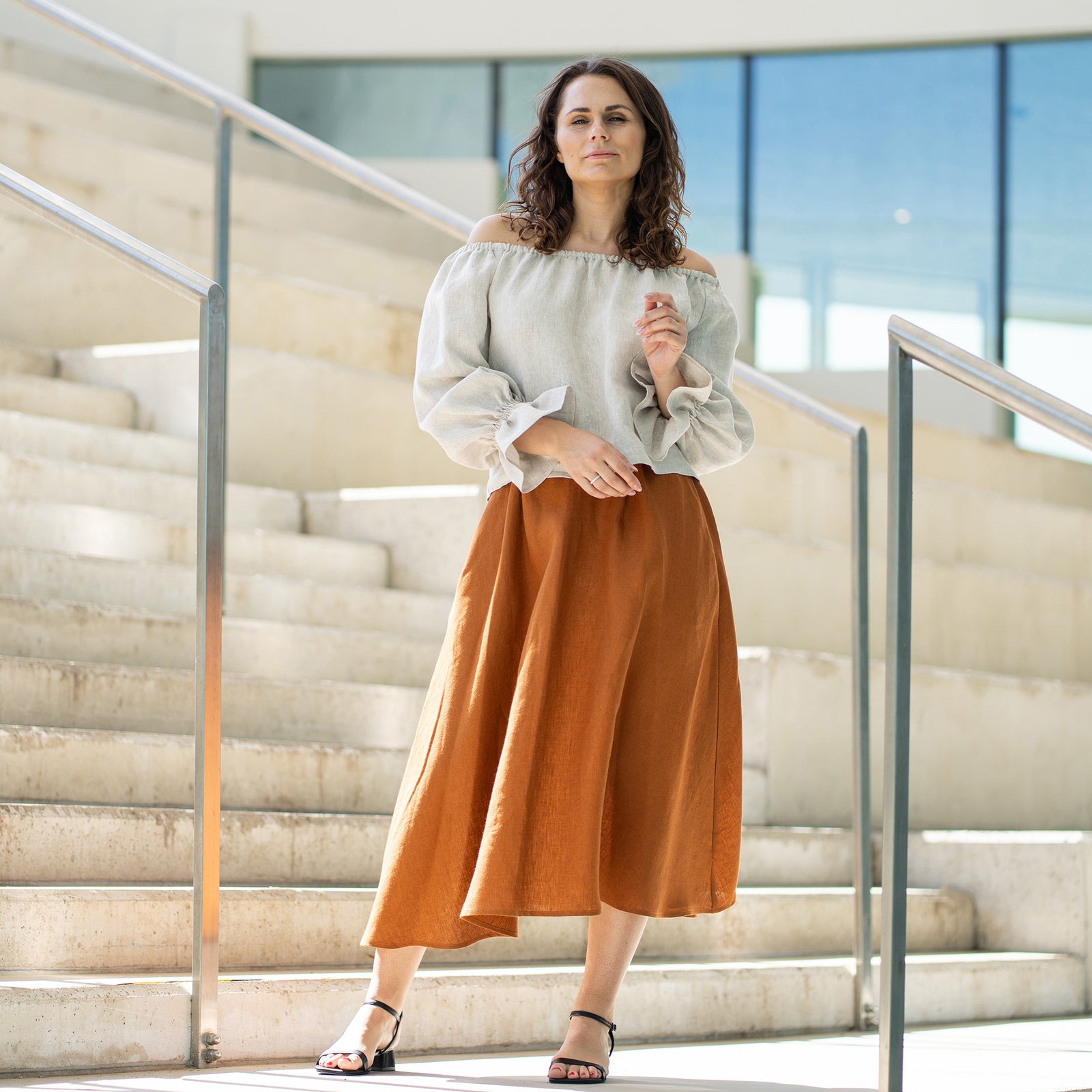 Een vrouw met bruin haar, gekleed in een lichtbeige top met blote schouders en pofmouwen en een lange roestbruine rok, poseert op betonnen trappen.