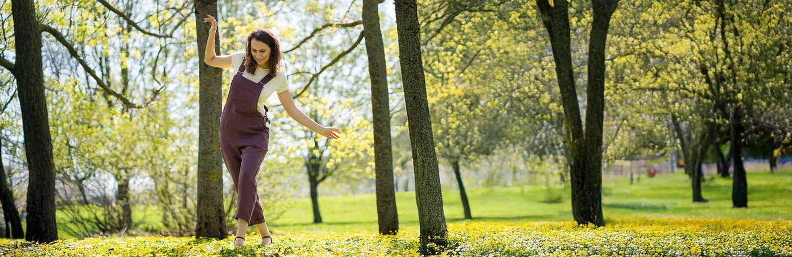 Woman wearing purple sleeveless jumpsuit layered over white t-shirt, relaxed fit women’s overalls style, breathable natural fabric one-piece outfit styled outdoors in spring nature setting