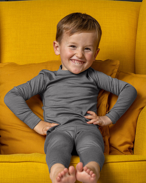 Cheerful boy in gray Merino wool base layer set sitting barefoot on a yellow armchair.