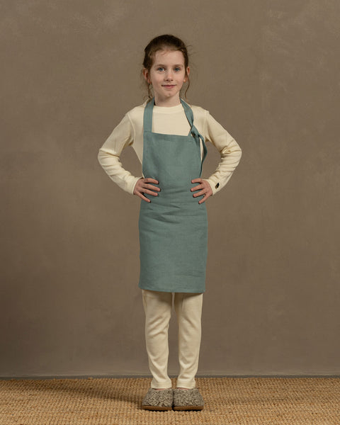 A young person with hair pulled back stands facing forward, wearing a long-sleeved cream-colored shirt, light-colored pants, and a mint green apron. Their hands are on their hips, and they are standing on a woven mat. The background is a solid, light brown color.