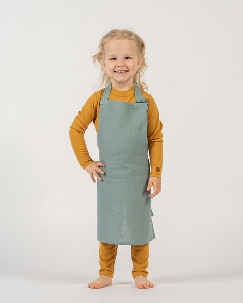 A young child with light curly hair, pulled into pigtails, stands facing forward with a wide smile. They are wearing a long-sleeved spicy yellow shirt and a mint green apron over it. The child has one hand on their hip. The background is white.