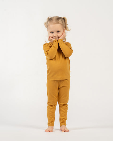 A cheerful child with curly blonde pigtails, wearing a yellow long-sleeved top and matching pants, stands barefoot on a white surface. The child is holding their hands to their cheeks and smiling at the camera.