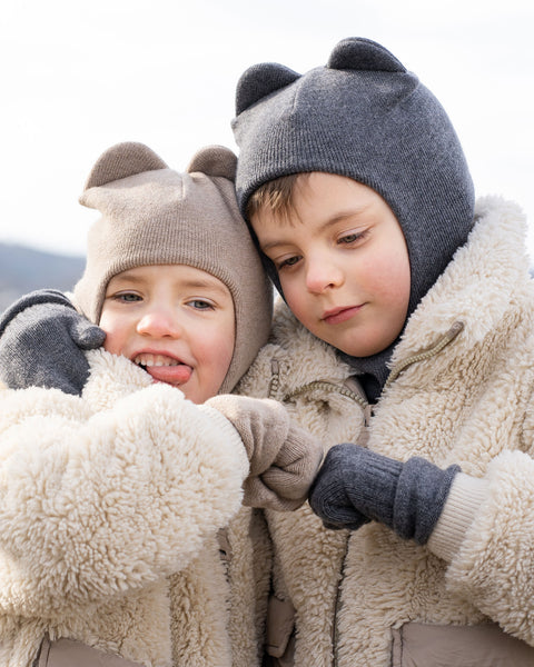 Two children, both wearing fuzzy, light-colored jackets and balaclavas with ear-like details, stand close together in an outdoor, possibly cold, setting. The child on the left wears a light brown balaclava and light gray gloves, looking towards the camera with a playful expression, tongue slightly out, and their gloved hand near their mouth.