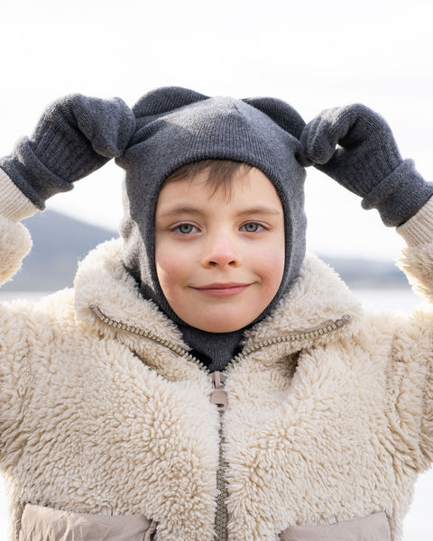 A child wearing a dark gray menique balaclava with two ear-like protrusions, gray gloves, and a light-colored, textured zip-up jacket over a gray turtleneck, stands outdoors. The child is looking directly at the camera with a slight smile and their gloved hands are raised to either side of their head, holding the top of the balaclava. The background is blurred, showing a light expanse, possibly water or ice, under a bright sky.