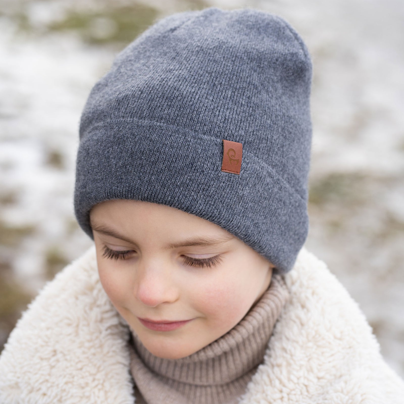 Een jong kind met kort bruin haar en een brede glimlach draagt winterkleding. Ze hebben een beige muts op, een beige coltrui en een crèmekleurig sherpa-jasje, met beige gebreide wanten. Op de achtergrond is een besneeuwd, wazig landschap te zien met wat lijkt op een wateroppervlak.