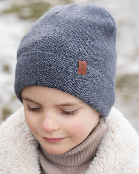 A close-up shot of a young child wearing a dark grey knit beanie with a brown label on the front, a beige turtleneck, and a cream-colored sherpa jacket. The child's eyes are cast downward, and the background is a blurred, lightly snowy outdoor scene.