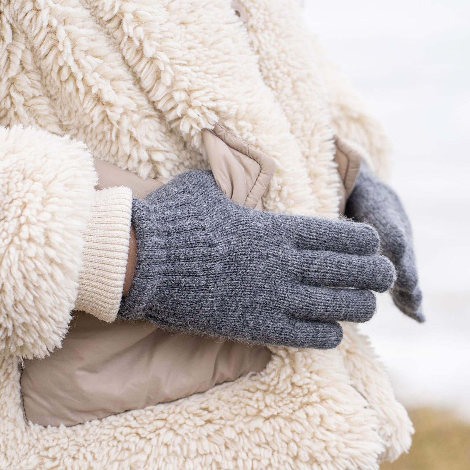 A hand wearing a light pink knitted glove with a ribbed wrist extends upwards and slightly to the left, revealing a plush, cream-colored sleeve on the arm, against a blurred light background.