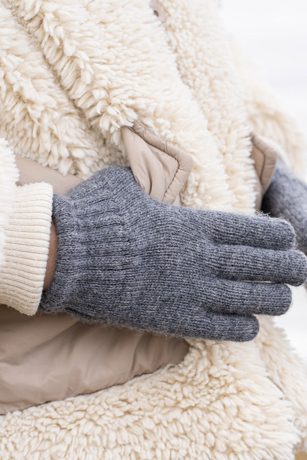 A person wearing a cream-colored sherpa coat with beige pockets and a light-colored ribbed cuff is putting on or adjusting a dark grey knitted glove. The background is a blurred, light outdoor scene.