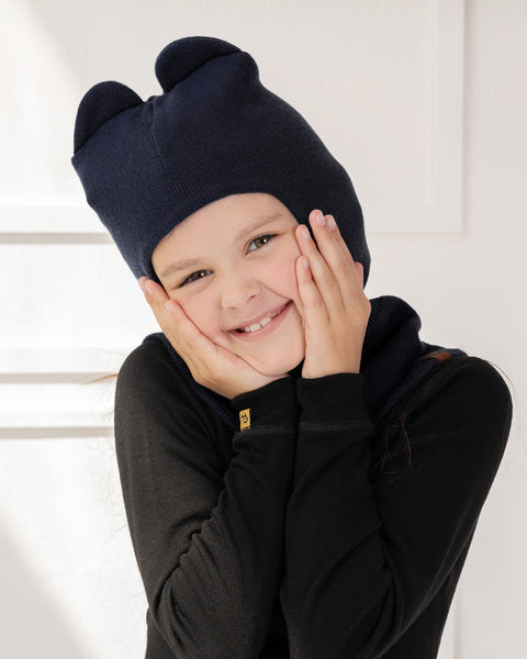 A girl holding her face with her palms, smiling, wearing the kids' cotton balaclava with playful ears in dark blue color.