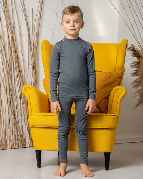 A boy standing in front of a yellow armchair, wearing the menique Kids' 160 Merino Pants in Perfect Grey color with a matching long sleeve top. 
