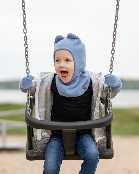A young child with blue eyes and an open-mouthed expression of excitement, is sitting in a playground swing. They are wearing a light blue balaclava with what looks like ears, light blue mittens, a grey speckled zip-up hoodie, and blue jeans. The background is a blurred outdoor scene with water and land.