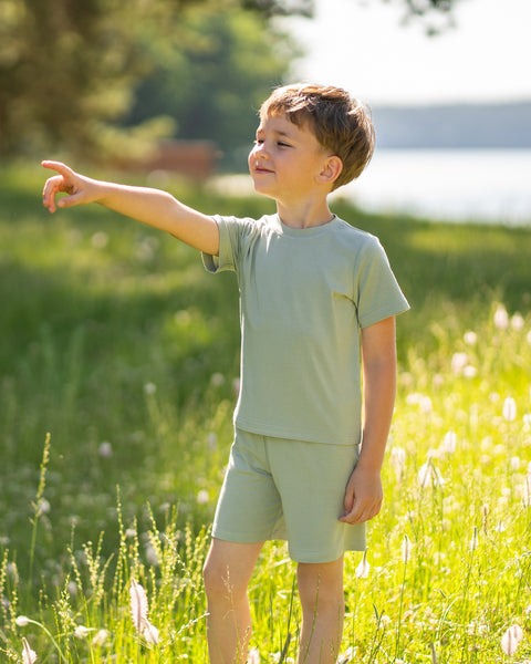 Young boy standing in a sunny meadow wearing a light green bamboo T-shirt and matching shorts. The lightweight kids' outfit is showcased in a natural outdoor setting, highlighting its soft, breathable comfort for everyday adventures.