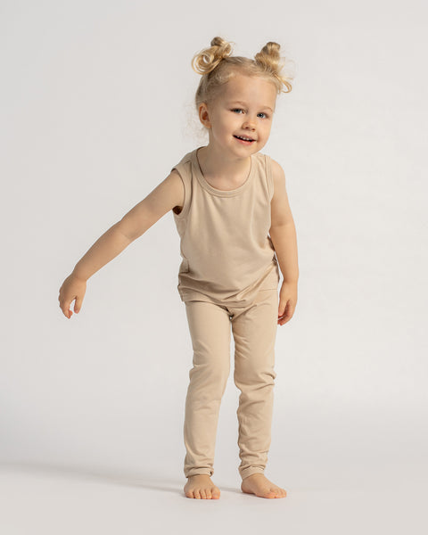 Happy child in a beige sleeveless outfit facing forward, smiling and holding hands up playfully against a neutral background.