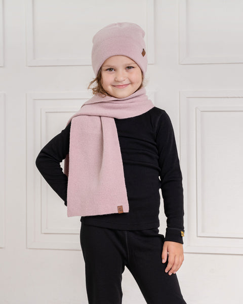 A child with wavy brown hair, wearing a dusty pink menique knit beanie with a small brown tag, a long pink knit scarf, a black long-sleeved shirt, and black pants, stands smiling at the camera. The child's right hand is on their hip, and their left arm is relaxed. The background is a white wall with decorative rectangular molding.