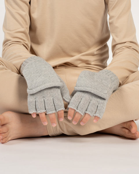 Child sitting cross-legged wearing light gray knitted convertible mittens, both mitten flaps open to reveal fingerless gloves.