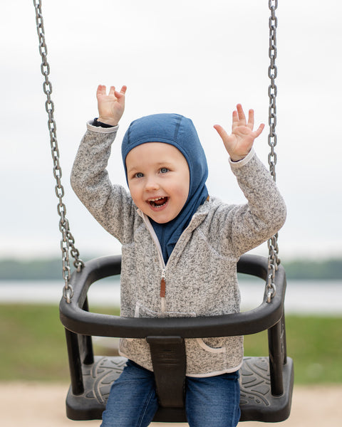 Toddler laughing with arms raised while swinging outdoors, wearing a hooded blue balaclava, a beige zip jacket, and blue jeans.