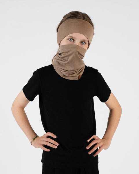 A studio portrait of a young girl standing with her hands on her hips, looking directly at the camera against a white background. She is wearing a black short-sleeved t-shirt, a beige headband, and a matching beige neck gaiter pulled up to cover her lower face and nose.