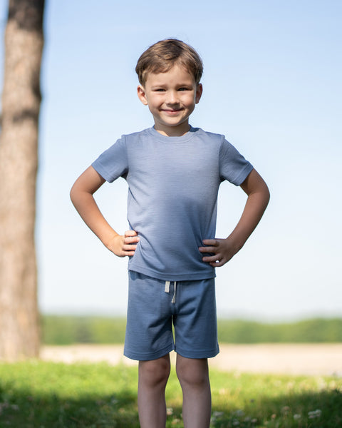 Young boy standing outdoors with his hands on his hips wearing a dusty blue Merino wool T-shirt and matching shorts. The lightweight, breathable kids' outfit is showcased in a sunny natural setting, offering soft comfort for everyday adventures and warm-weather play.