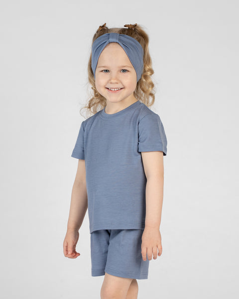 A young girl with blonde hair styled in two pigtails smiles at the camera. She is wearing a dusty blue merino short-sleeve t-shirt, matching dusty blue merino shorts, and a matching dusty blue merino ribbon headband. She is standing against a plain light gray background.