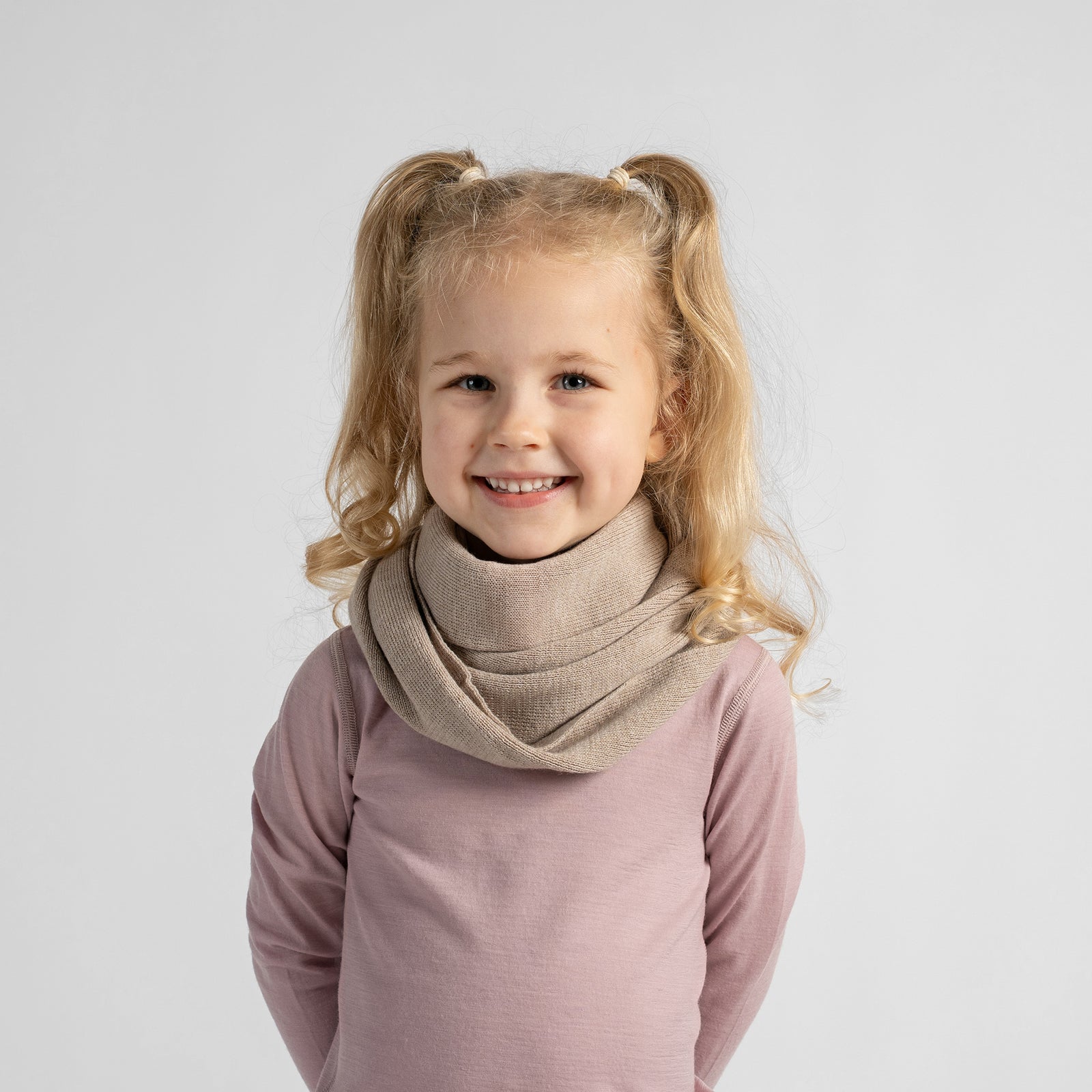 A young girl with blonde hair smiling, wearing a soft beige knit cowl neck warmer over a dusty pink long-sleeve top, photographed against a solid light gray background.