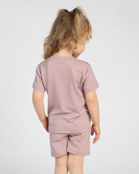 A young child is shown from behind, wearing a dusty pink short-sleeve t-shirt and matching shorts. Their blonde hair is styled in a loose half-up ponytail, and they are standing against a light background.