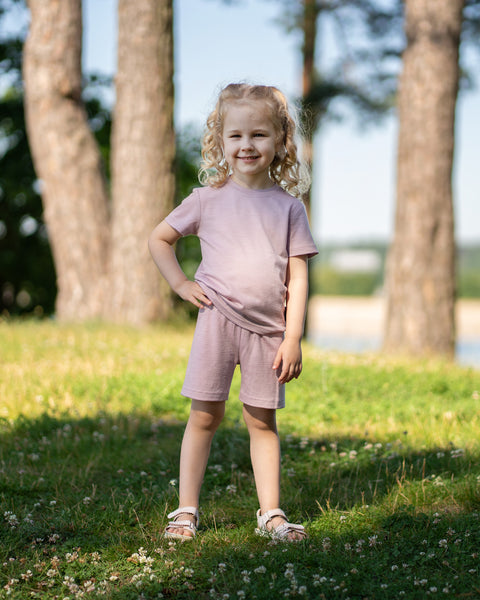 Young girl standing in a sunny park wearing a dusty pink Merino wool T-shirt and matching shorts. The lightweight, breathable kids' outfit is showcased in a natural outdoor setting, offering soft comfort for everyday play and warm-weather adventures.