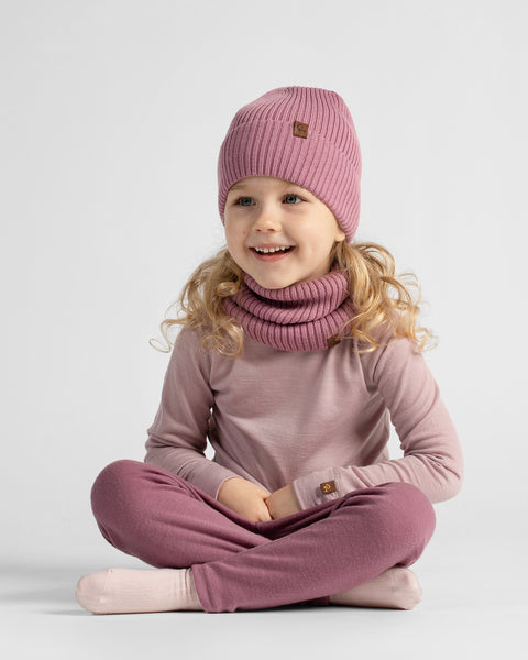 A young girl sitting cross-legged against a white backdrop, wearing a dusty rose loungewear outfit with a matching pink beanie and neck scarf.