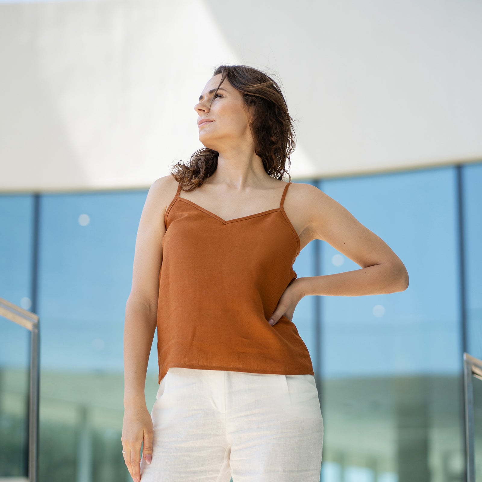 Een vrouw met bruin haar draagt een amandelbruine top met V-hals en witte broek, poserend binnen voor grote glazen ramen.