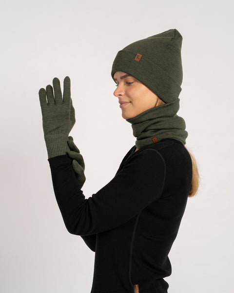 Woman wearing a dark green knit beanie, matching gloves, and a neck gaiter, dressed in a black long-sleeve top, raising one gloved hand in profile view against a plain background.