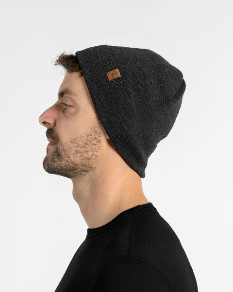 A man with light skin, blue eyes, and a beard. He wears a dark gray colored menique knit beanie with a small brown patch on the side, and a black crew-neck shirt.