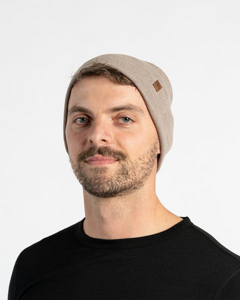 A man with light skin, blue eyes, and a beard smiles faintly while looking forward. He wears a creamy beige colored menique knit beanie with a small brown patch on the side, and a black crew-neck shirt.