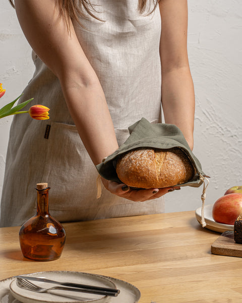 Een persoon in een linnen schort houdt een brood op een houten tafel met andere etenswaren.